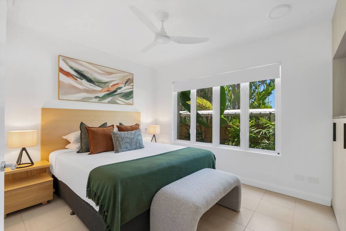 Ein modernes Schlafzimmer mit einem großen Bett, einem Deckenventilator, einem Fenster mit Blick auf Grünflächen und einem Gemälde an der Wand.