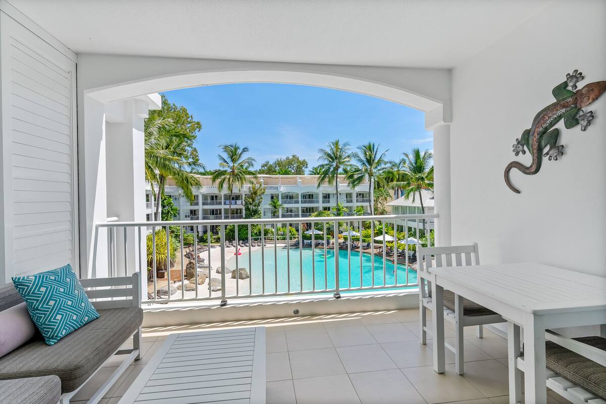 Ein Balkon mit Blick auf einen Swimmingpool, Palmen und ein Gebäude im Hintergrund.