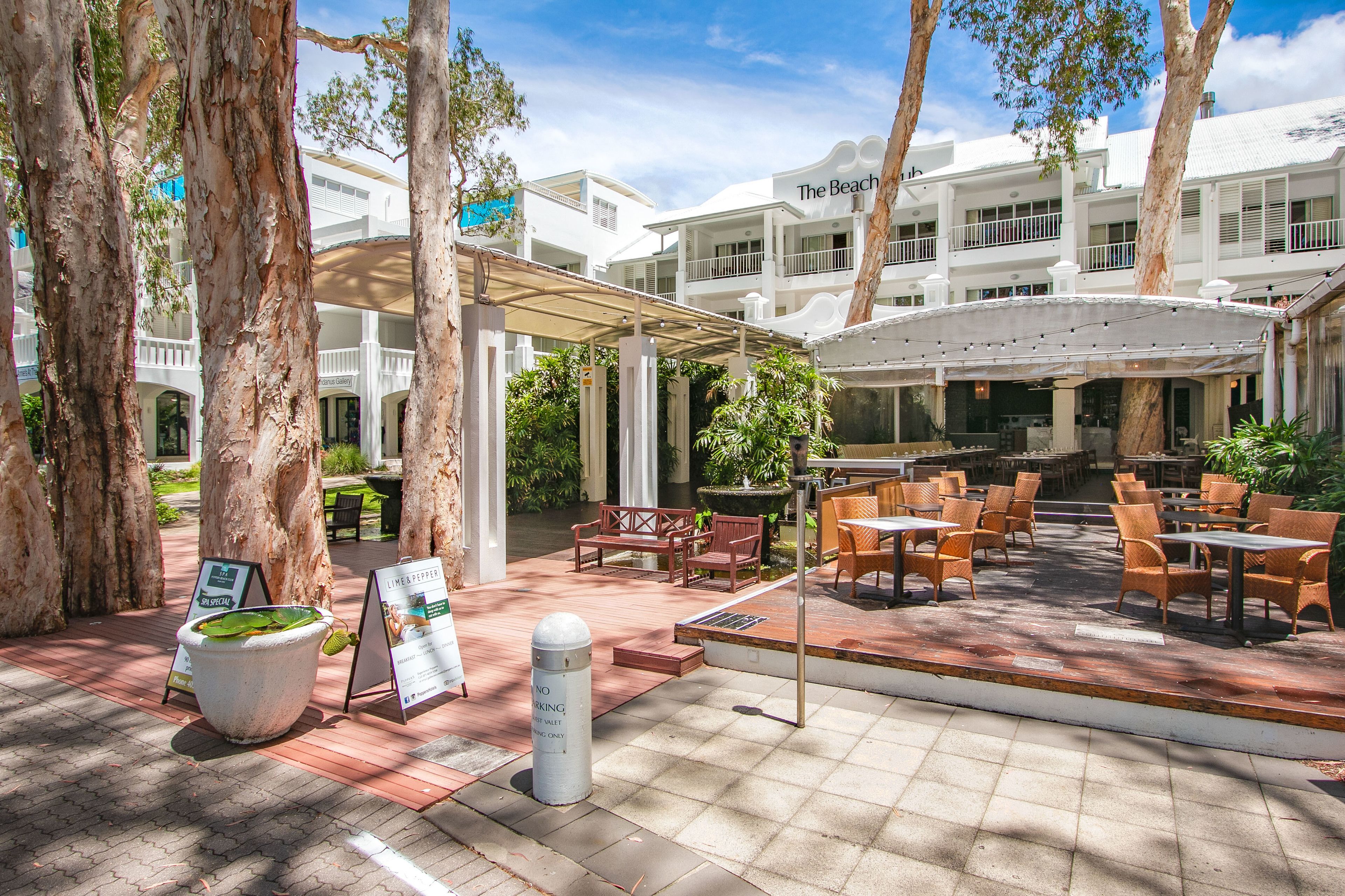 A covered outdoor seating area with tables and chairs, surrounded by trees and a building with balconies.