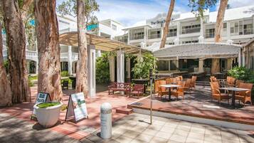 A covered outdoor seating area with tables and chairs, surrounded by trees and a building with balconies.