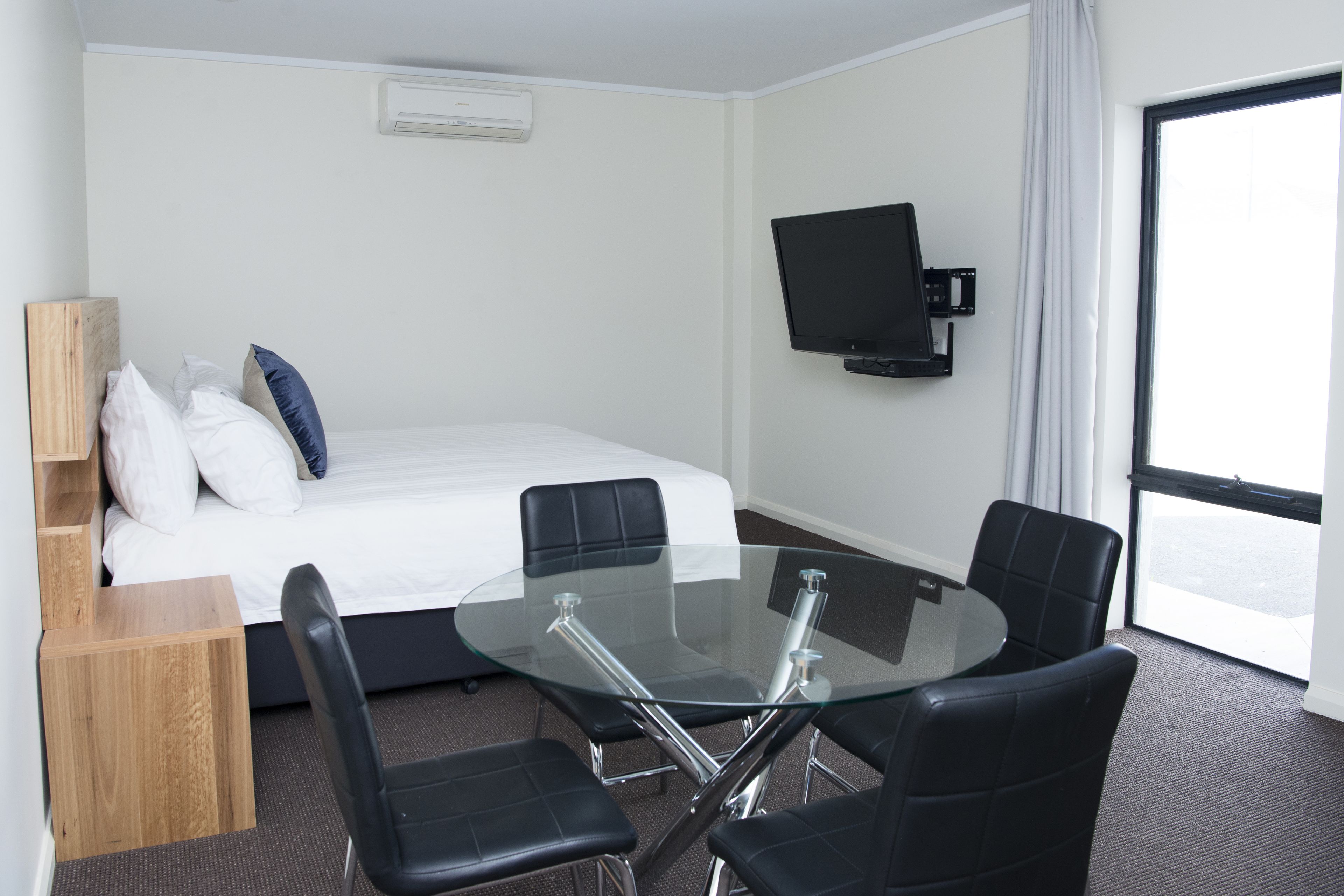 A hotel room with a bed, a glass table with four chairs, a flat-screen TV mounted on the wall, and a large window.