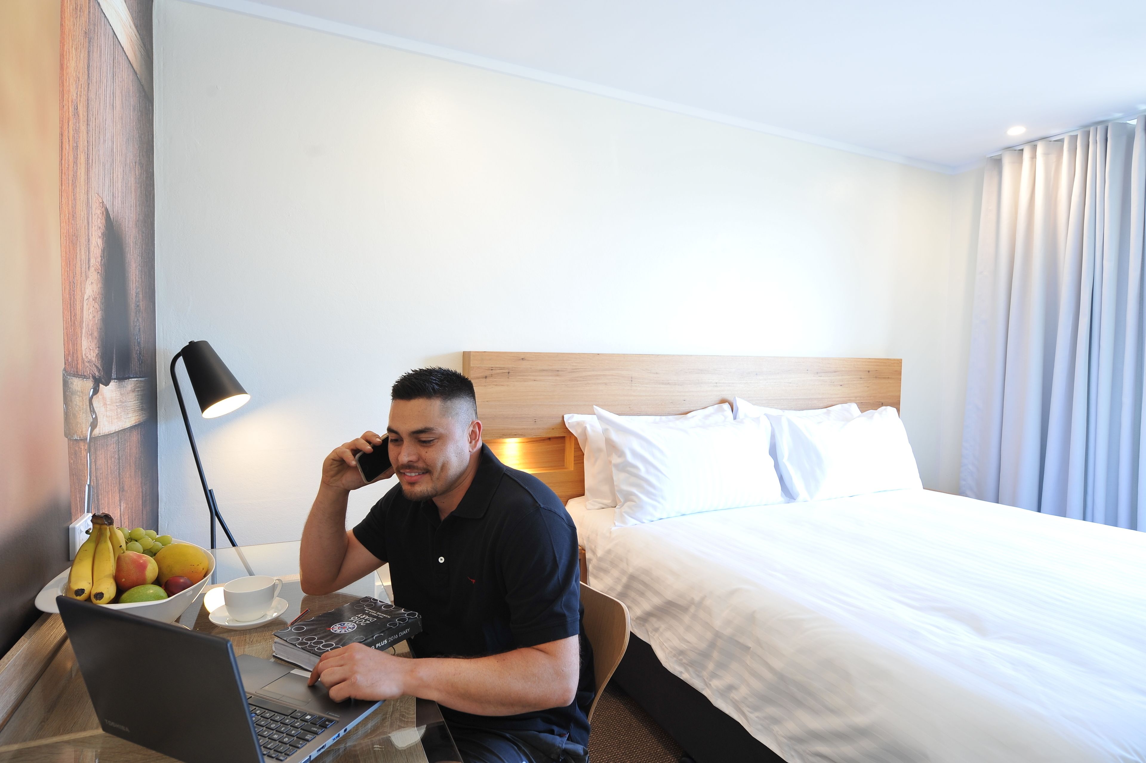 A hotel room with a bed, a desk with a laptop, a fruit bowl, and a lamp.