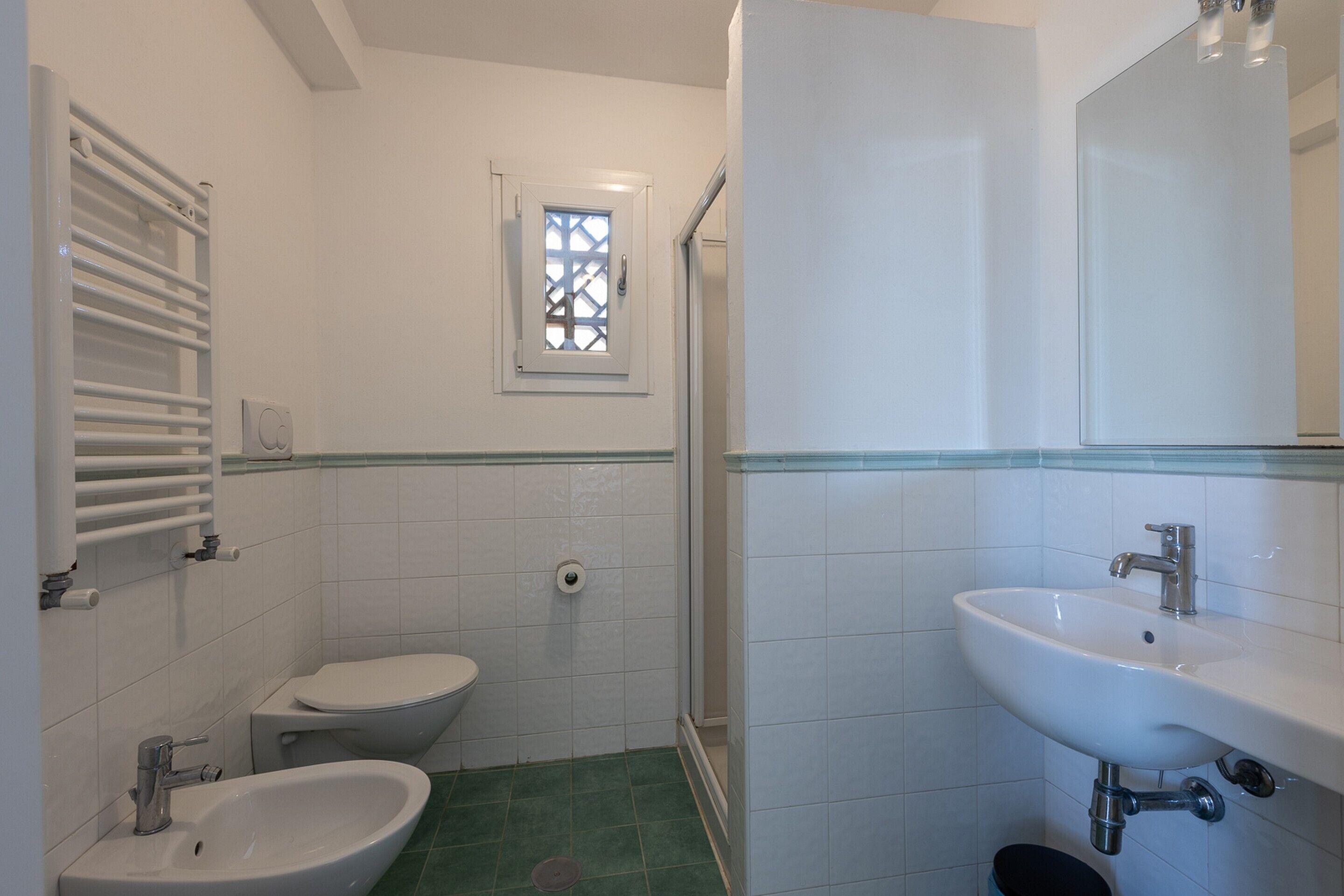 Une salle de bain comprenant des toilettes, un bidet, un lavabo et une fenêtre ornée d’un motif en treillis.