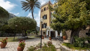 Pátio com um edifício, janelas em arco e varanda, cercado por plantas em vasos e área de estar ao ar livre.