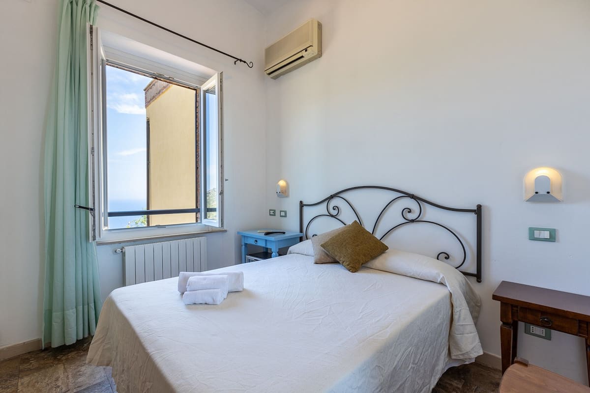 A neatly made bed with pillows and a set of towels, a small bedside table, a nightstand, and a window with curtains.