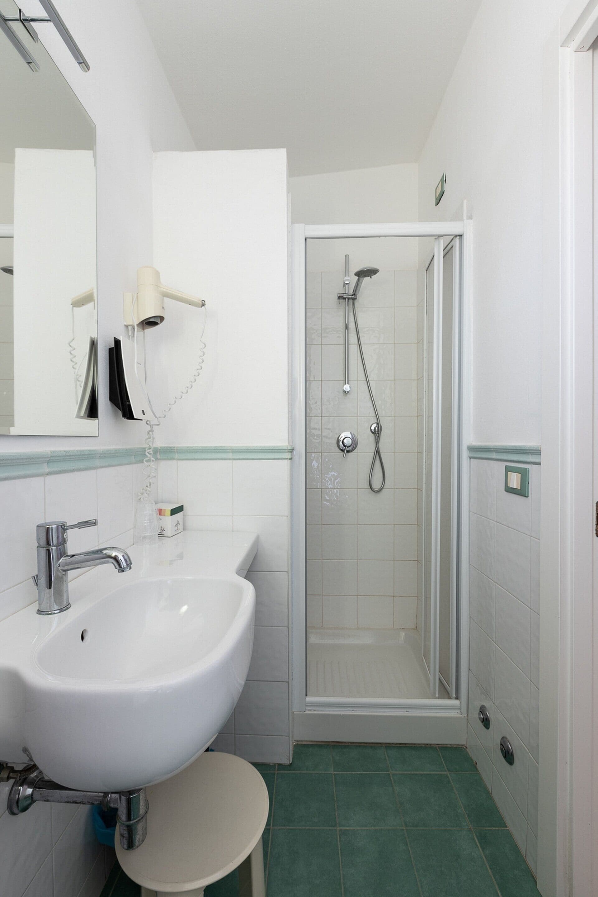 Ein Badezimmer mit Waschbecken in Weiß, einem Spiegel, einer Dusche mit Glastür und grün gefliestem Boden.