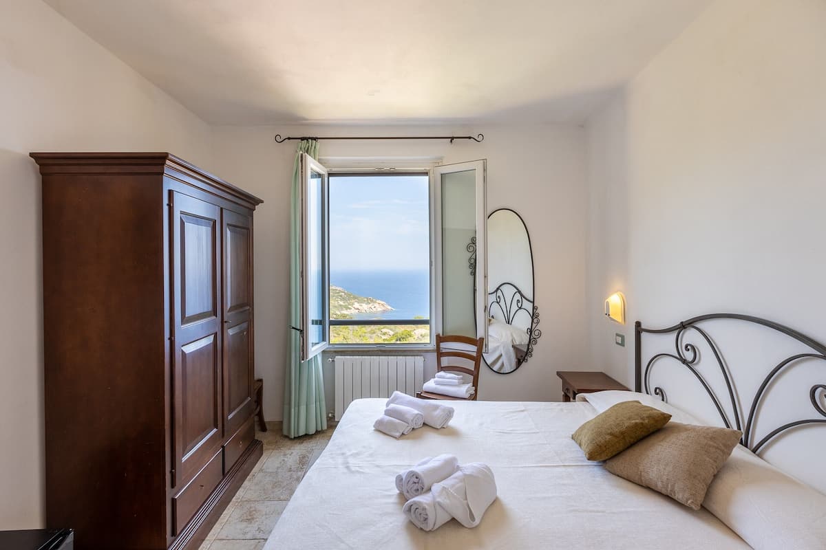 A bedroom with a bed, a wardrobe, a chair, a nightstand, a mirror, and a view of the sea.
