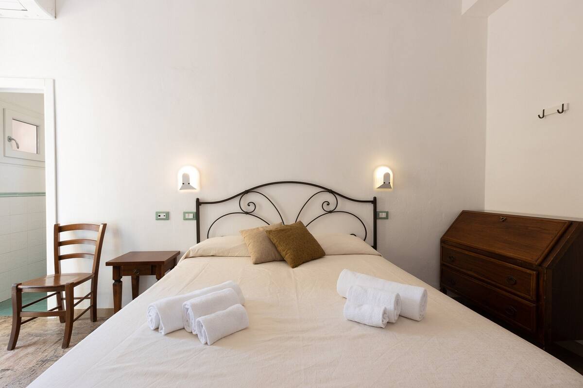 A neatly made bed with pillows, a chair, a small table, and a wooden dresser in a simple room with two wall-mounted lights.