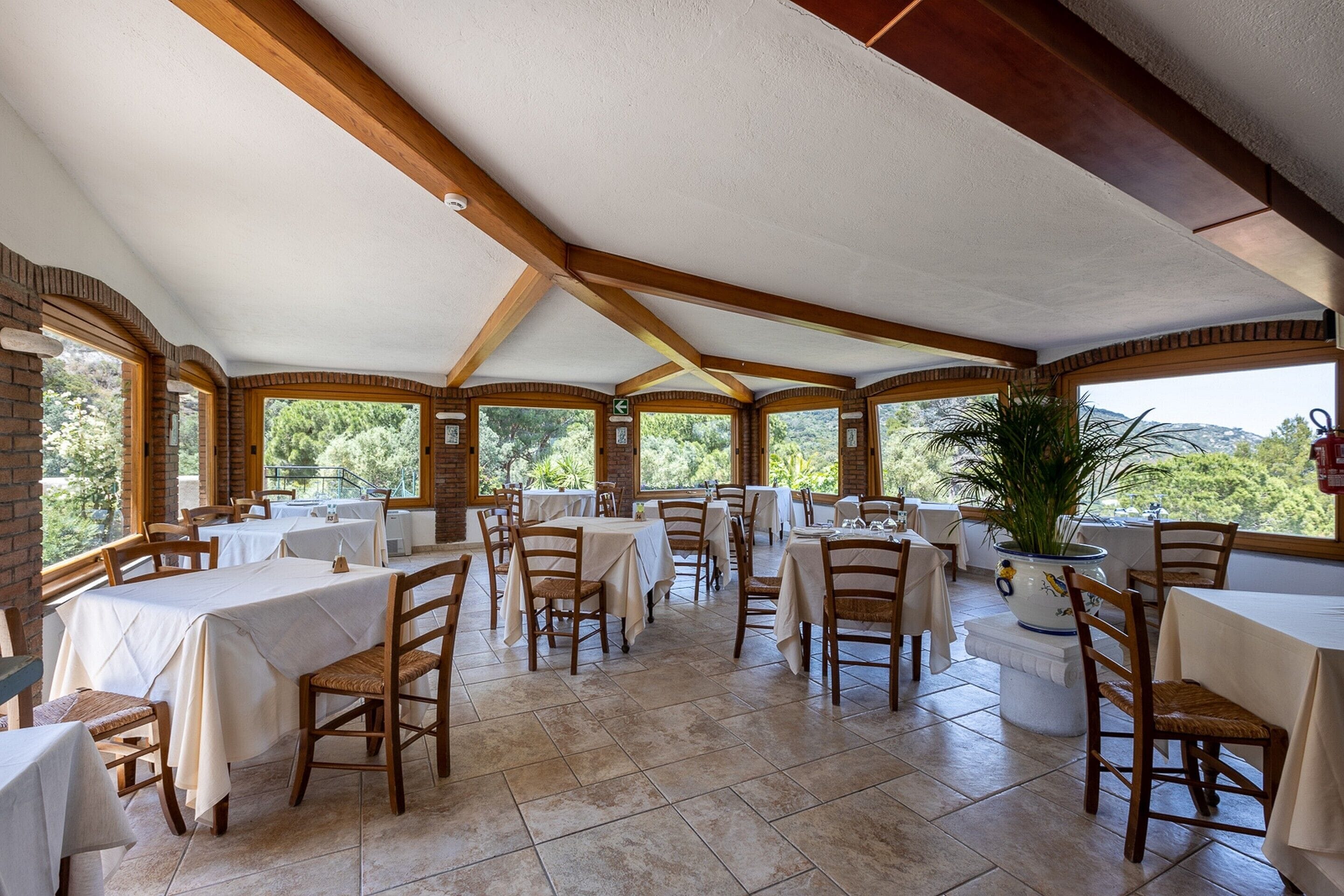 A spacious indoor dining area with multiple tables and chairs, large windows, and a view of greenery outside.