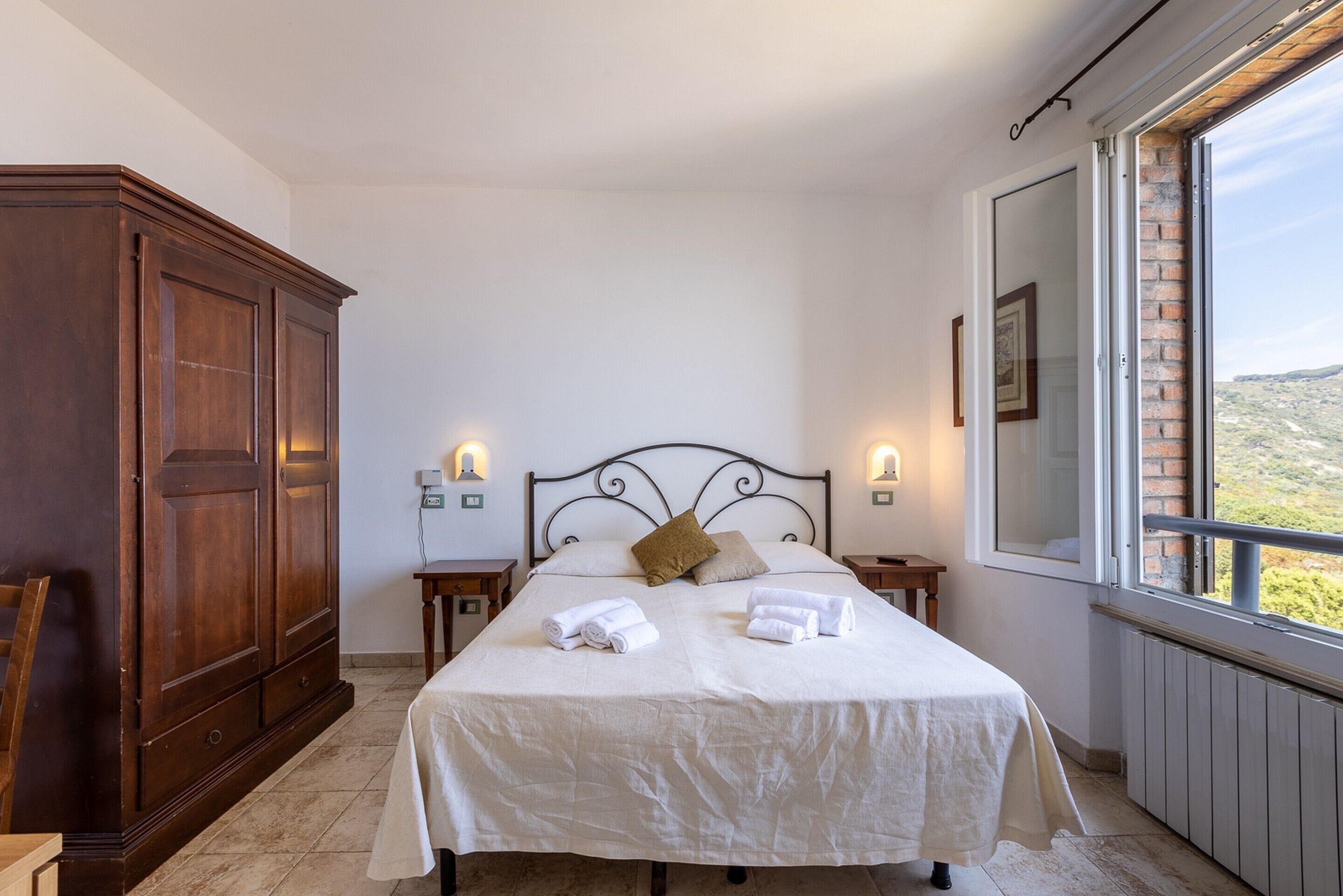 Un dormitorio con un amplio armario de madera, una cama con ropa de cama blanca, dos mesitas de noche con lámparas y vistas a través de una ventana.