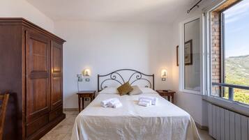 Un dormitorio con un amplio armario de madera, una cama con ropa de cama blanca, dos mesitas de noche con lámparas y vistas a través de una ventana.