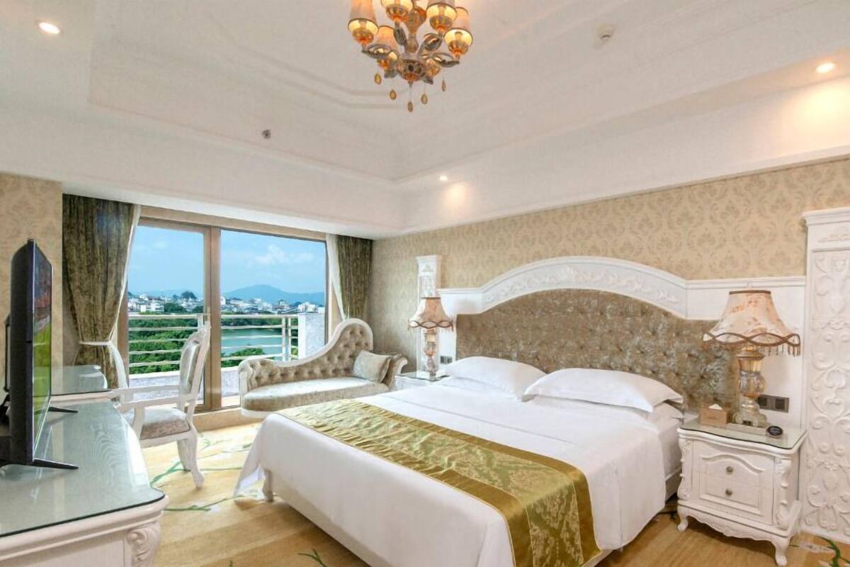 A hotel bedroom with a large bed, a television, a balcony with a view, and a chandelier.