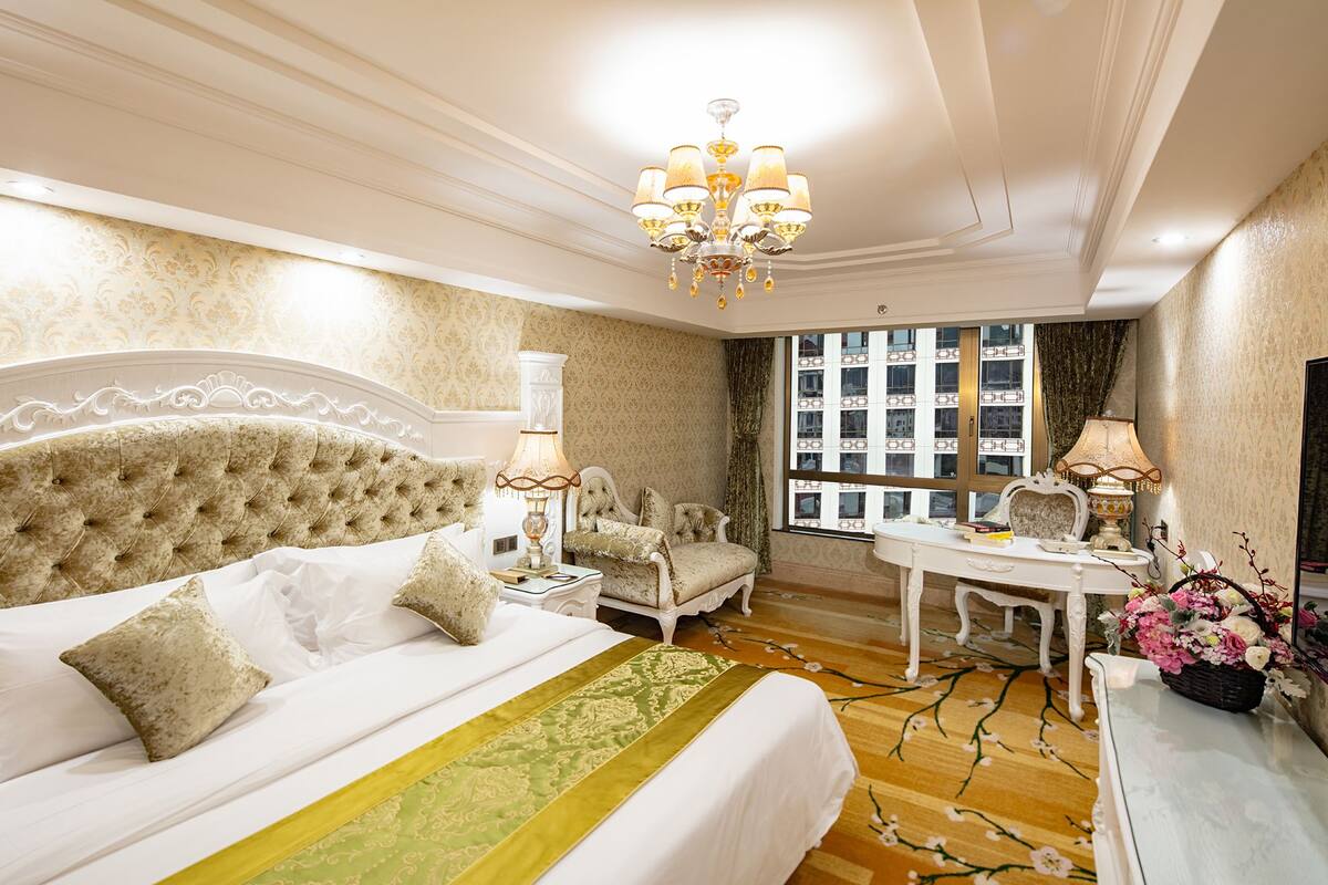 A luxurious bedroom with a large bed, a chandelier, a desk, and a view of the city.