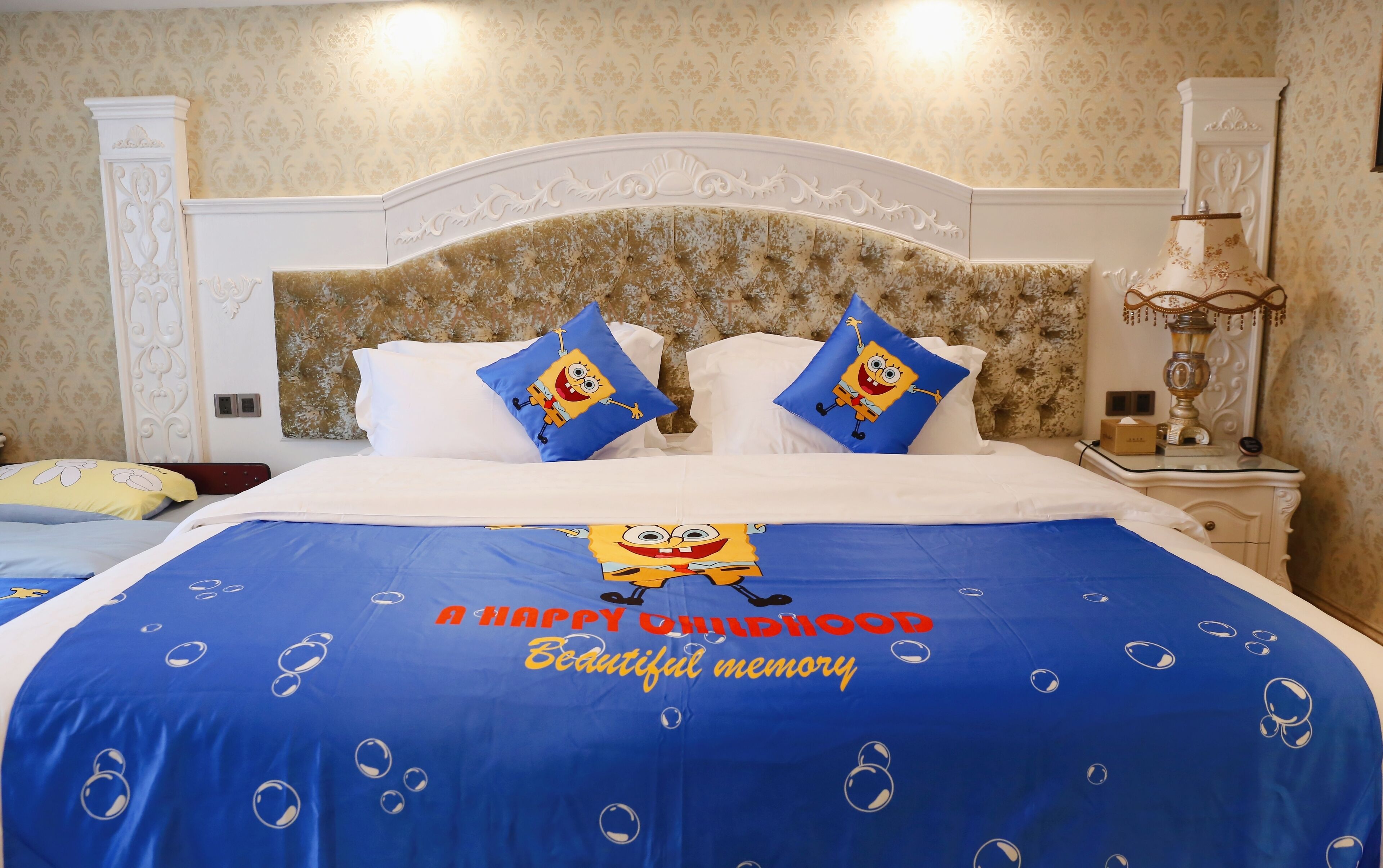 Una habitación de hotel con una cama que incluye una colcha azul con diseño de Bob Esponja y almohadas, un cabecero blanco y una mesita de noche con una lámpara.