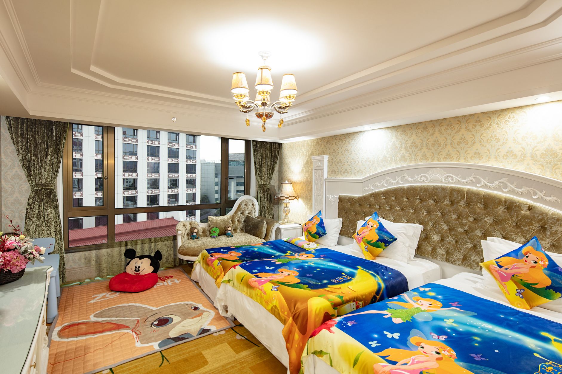 A hotel room with two beds, a large window with a city view, a chandelier, and a Mickey Mouse plush toy on the floor.
