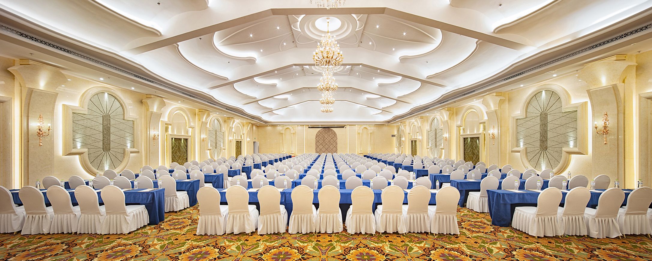 A large conference room with rows of tables and chairs, a patterned carpet, and a chandelier.