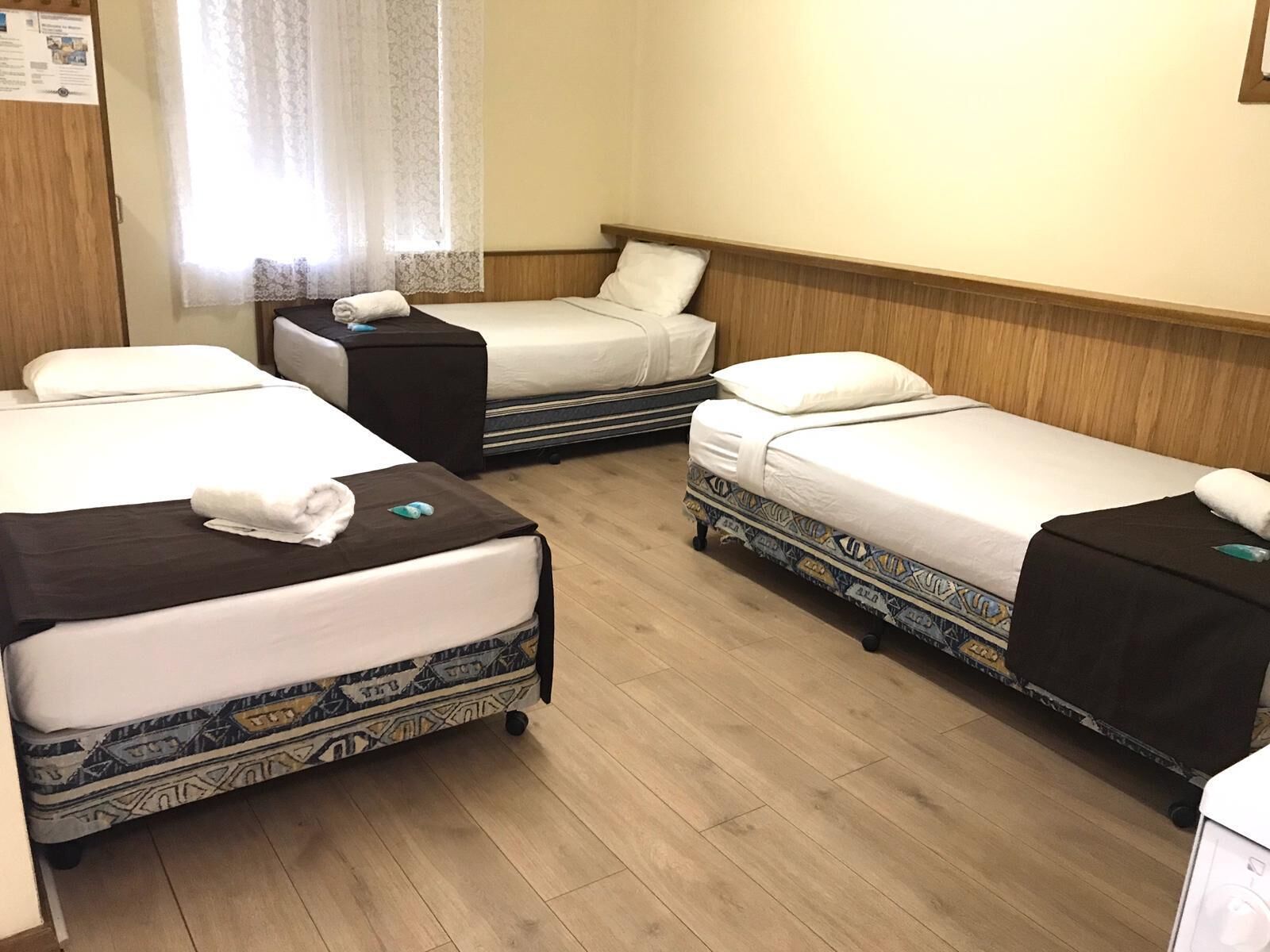 Een hotelkamer met vier eenpersoonsbedden, elk met een kussen en een opgevouwen handdoek. De kamer heeft houten vloeren en een houten hoofdbord.
