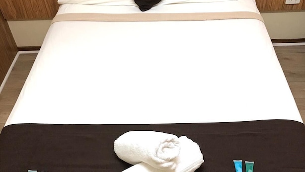 A neatly made bed with a black pillow, a white pillow, and a white towel folded on the bed. There are two small tubes of cream or lotion on the bed. - Angel's Rest Motel (Moree)