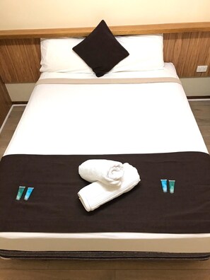 Un lit bien fait, avec un oreiller noir, un oreiller blanc et une serviette blanche pliée posée sur le lit. On peut y voir deux petits tubes de crème ou de lotion.