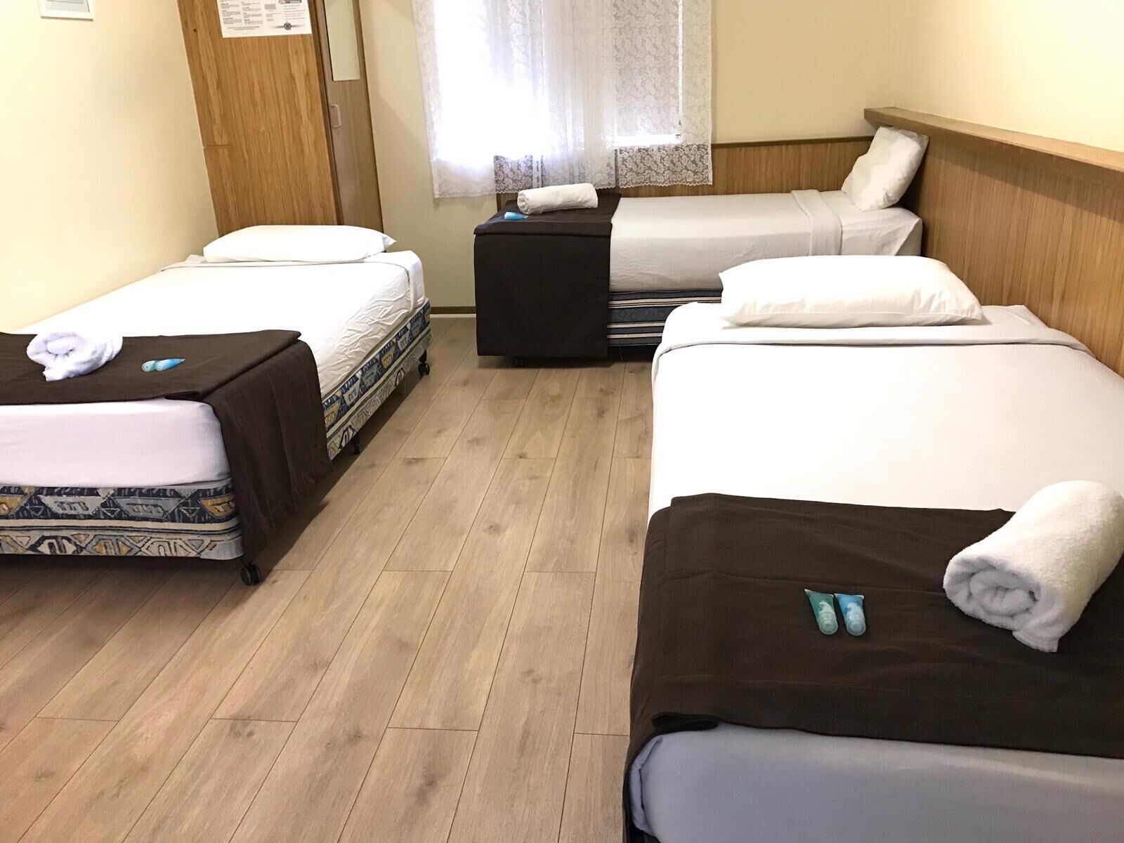 Habitación de hotel con tres camas alineadas, piso de madera y ventana con cortinas de encaje.