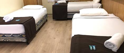 Habitación de hotel con tres camas alineadas, piso de madera y ventana con cortinas de encaje.