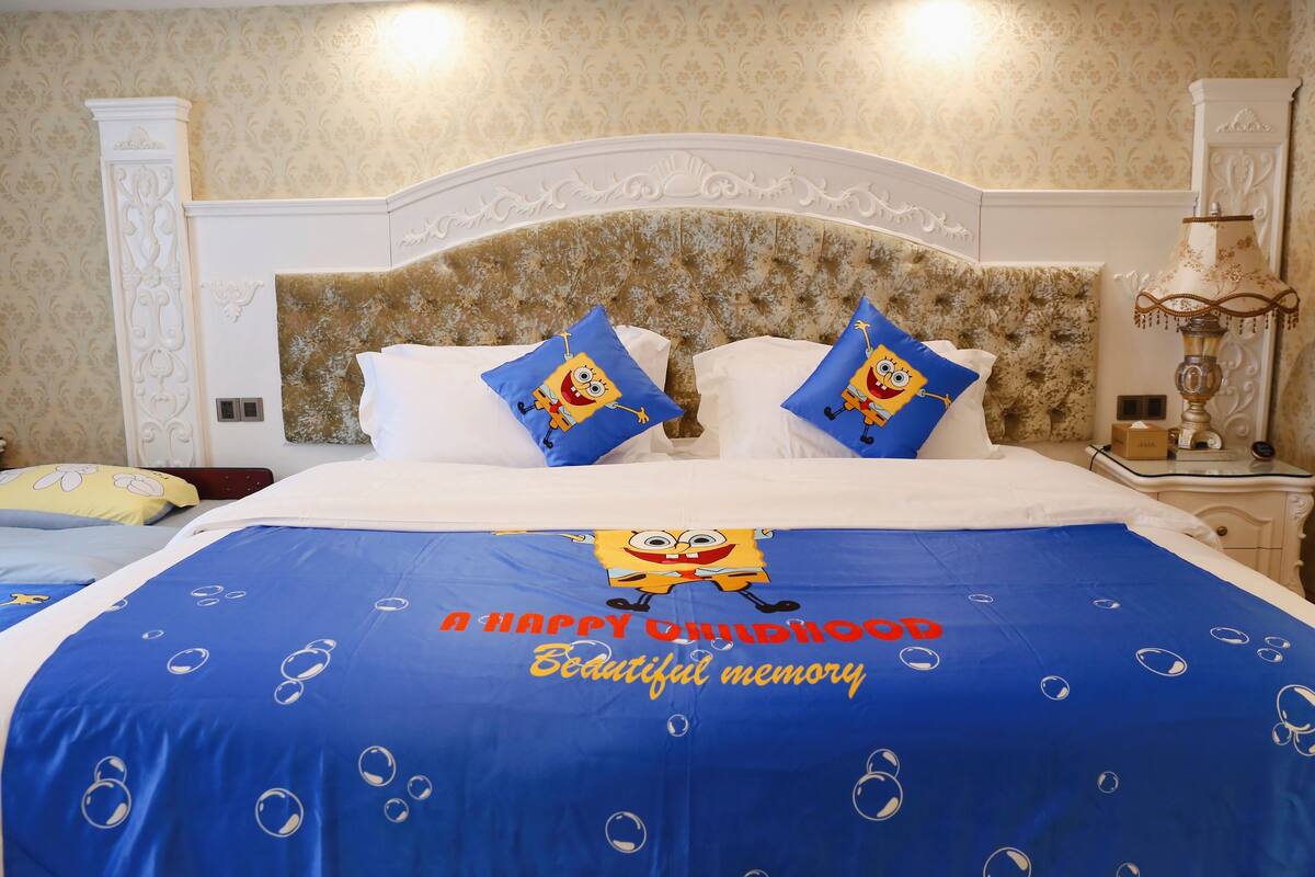 A hotel room with a bed featuring a blue bedspread with a SpongeBob SquarePants design and pillows, a white headboard, and a nightstand with a lamp.