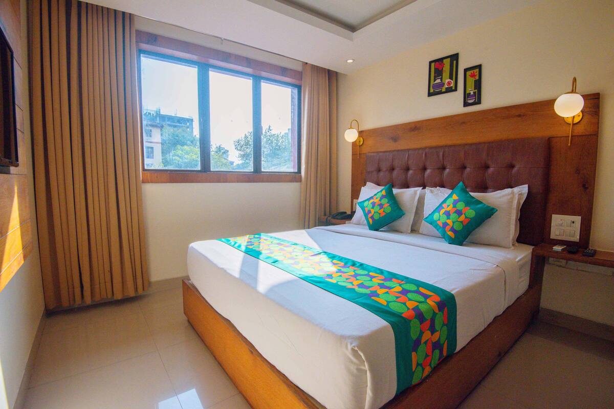 A hotel room with a large bed, a window with curtains, a headboard with wall-mounted lights, and a bedside table with a lamp.