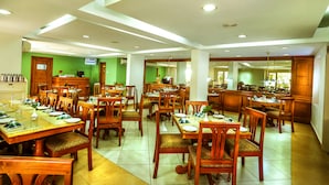 A spacious dining area with multiple wooden tables and chairs, a kitchen in the background, and a green wall. - Krishna Inn (Chavakkad)