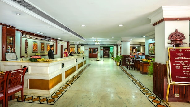 A hotel reception area with a reception desk, chairs, a wall-mounted clock, and a signboard with restaurant timings. - Krishna Inn (Chavakkad)