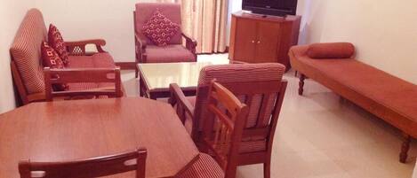 Una sala de estar con una mesa de comedor de madera, dos sillas, un sofá, un televisor sobre un mueble bajo y una cortina estampada.