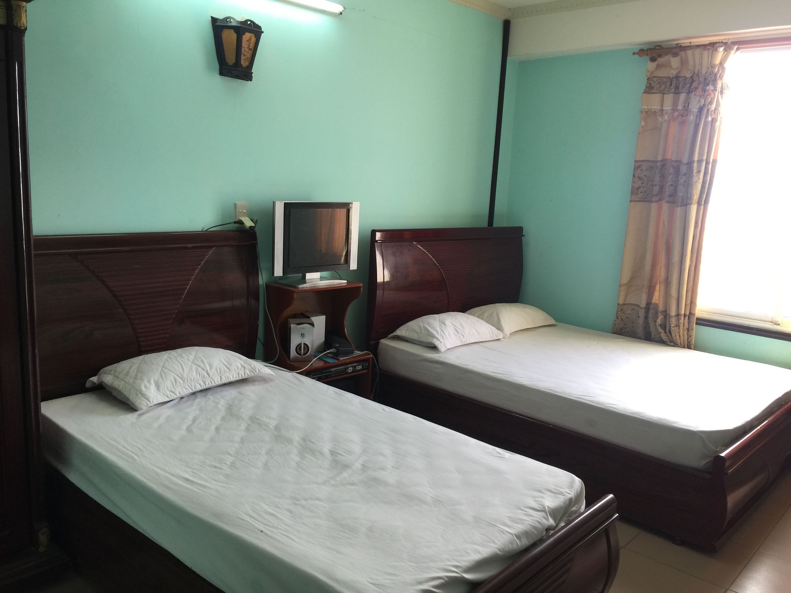 A room with two single beds, a desk with a computer, a wall-mounted lamp, and a window with curtains.