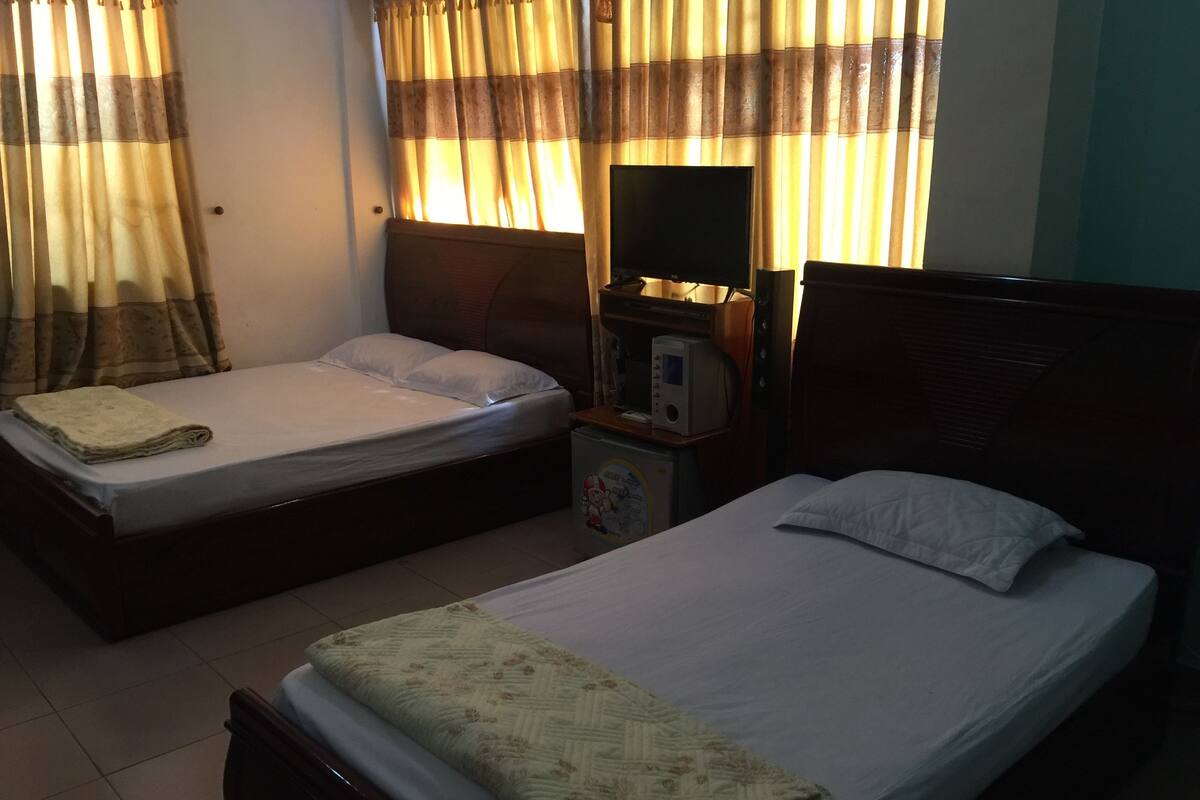 A room with two single beds, each with a pillow and a folded blanket. There is a small table with a microwave and a coffee maker. The room has a window with curtains, and the walls are plain.