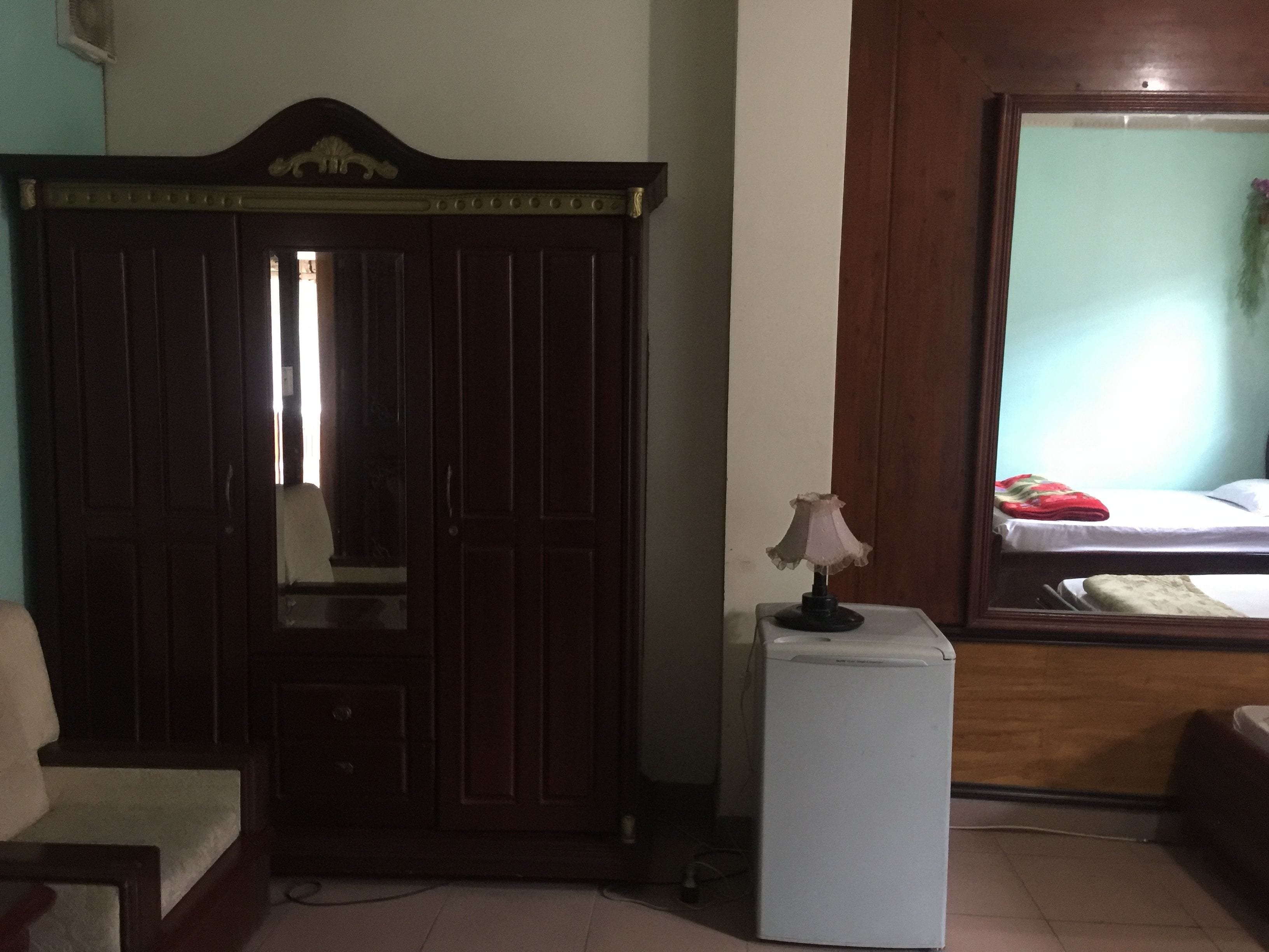 A bedroom with a wardrobe, a small table with a lamp, a bed, and a mirror.
