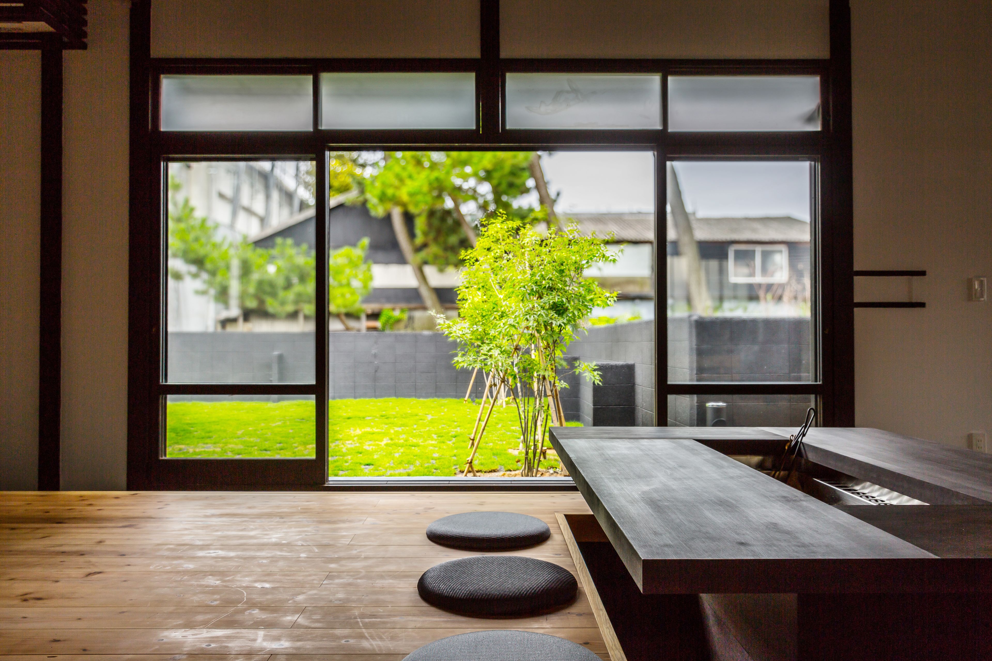 Een modern interieur met een houten vloer, een eettafel met krukken, en uitzicht door grote glazen deuren op een groen buitengebied met een boom.