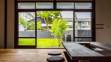 Een modern interieur met een houten vloer, een eettafel met krukken, en uitzicht door grote glazen deuren op een groen buitengebied met een boom.