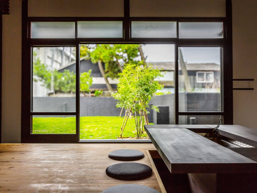Een modern interieur met een houten vloer, een eettafel met krukken, en uitzicht door grote glazen deuren op een groen buitengebied met een boom.
