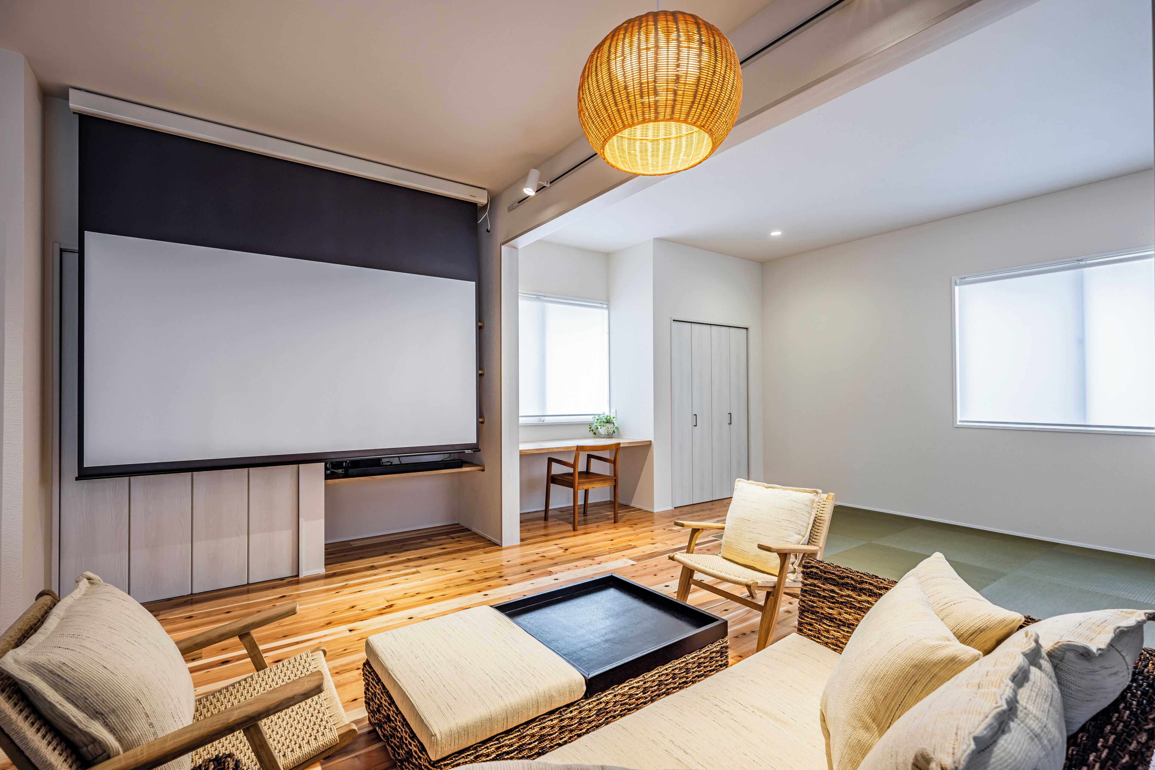 A modern living room with a flat-screen TV, wooden flooring, wicker furniture, and a large window.