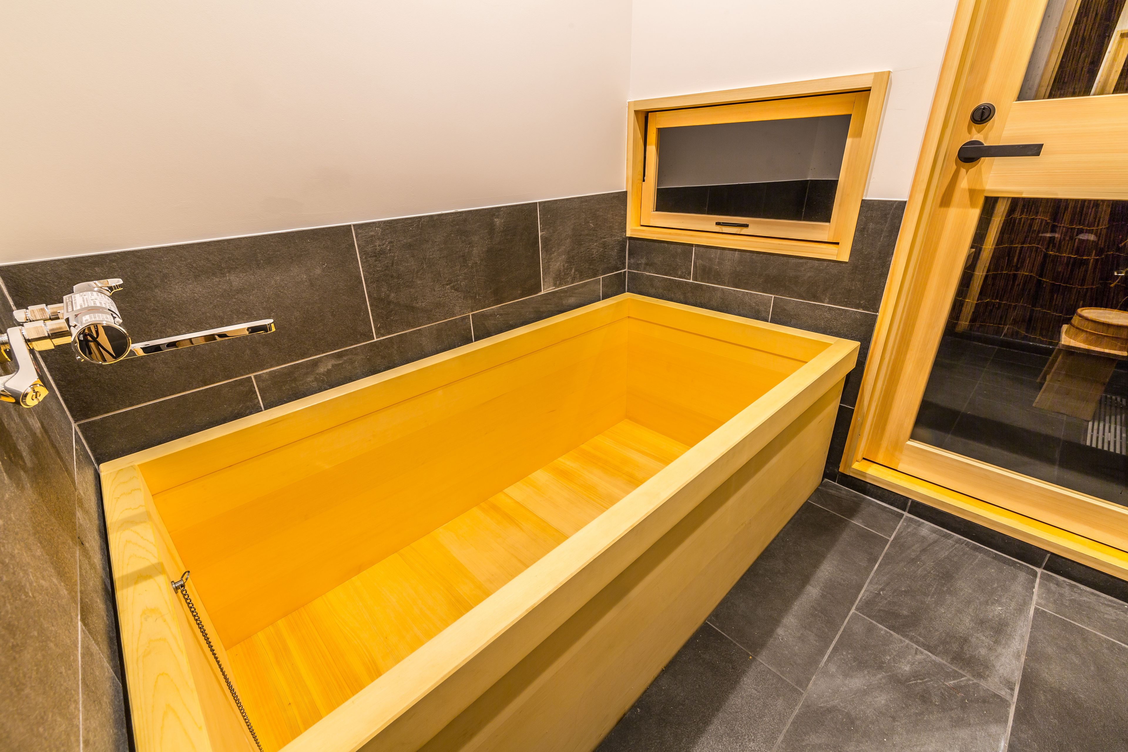 A modern bathroom with a wooden bathtub, a glass shower enclosure, and a small window above the bathtub.