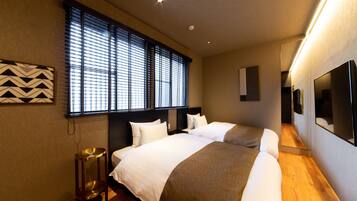 Een hotelkamer met twee bedden, een groot raam met zonwering, een nachtkastje en een aan de muur bevestigde televisie.