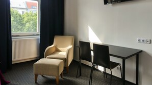 Et hotelværelse med seng, et vindue med gardiner, et fjernsyn monteret på væggen, et skrivebord med to stole og en radiator.