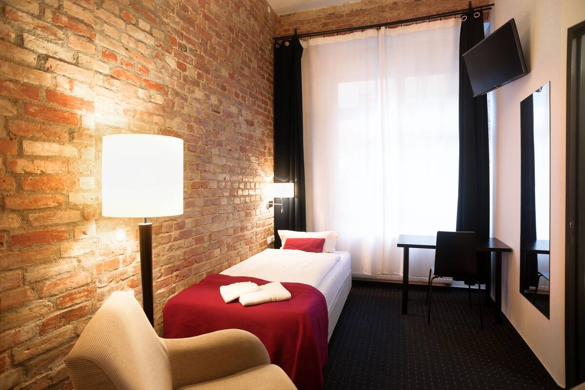 A hotel room with a brick wall, a bed with a red bedspread, a desk with a chair, a flat-screen TV mounted on the wall, and a window with sheer curtains.