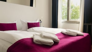 En pænt redt seng med hvidt og pink sengetøj, en senelampe og et vindue med gardiner.