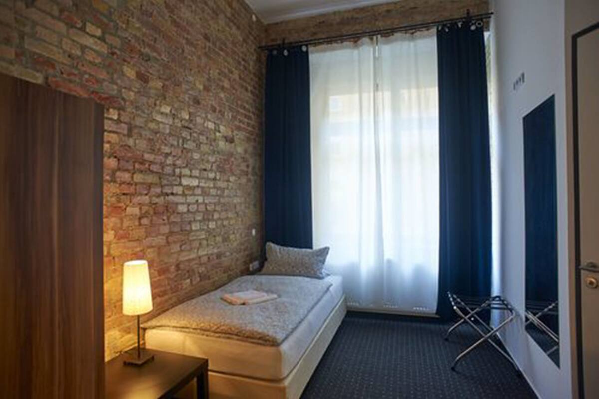 A small, cozy room with a brick wall, a single bed, a bedside lamp, a window with curtains, and a door.