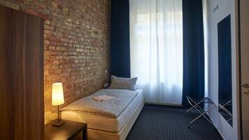 A small, cozy room with a brick wall, a single bed, a bedside lamp, a window with curtains, and a door.