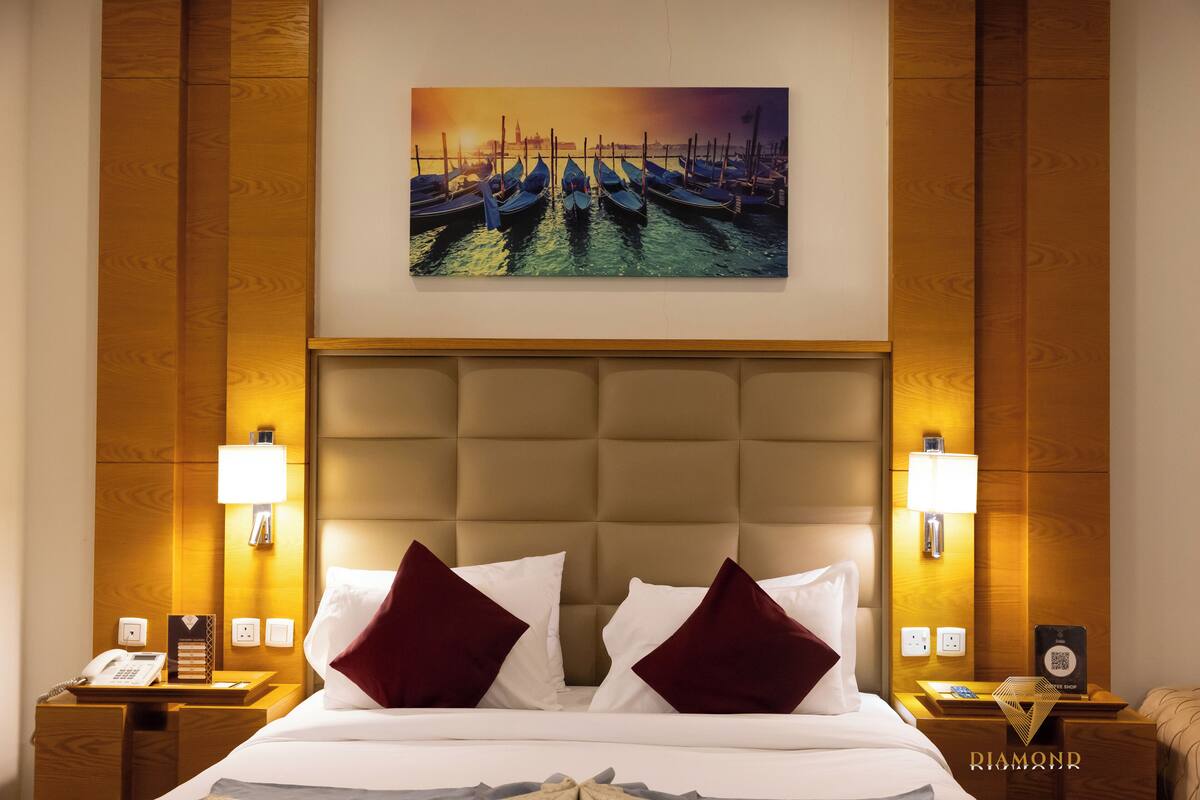 A hotel room with a large bed, two nightstands, a painting of boats, and a wooden headboard.