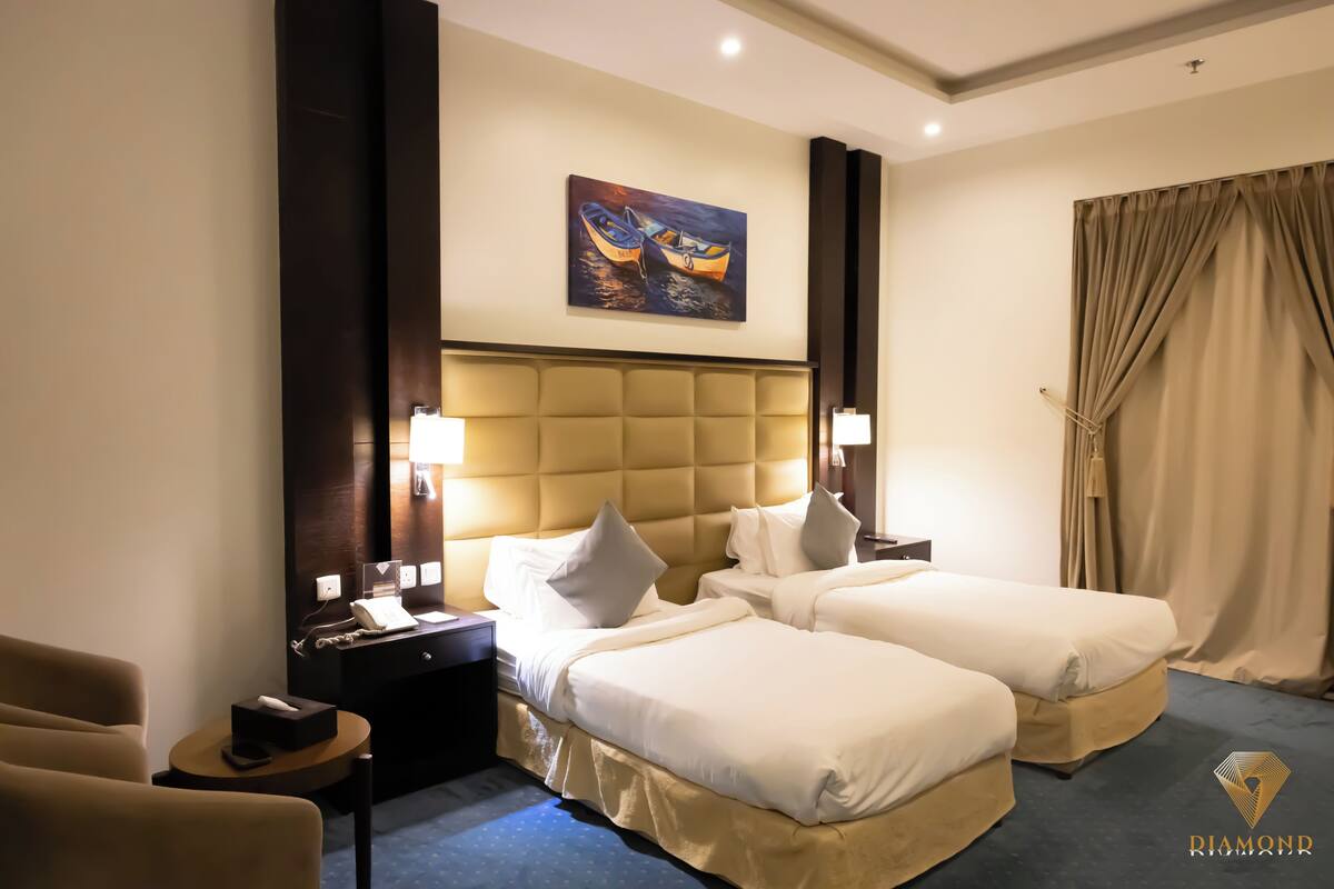 A hotel room with two beds, a headboard, a painting of boats, a desk with a phone, and a chair.