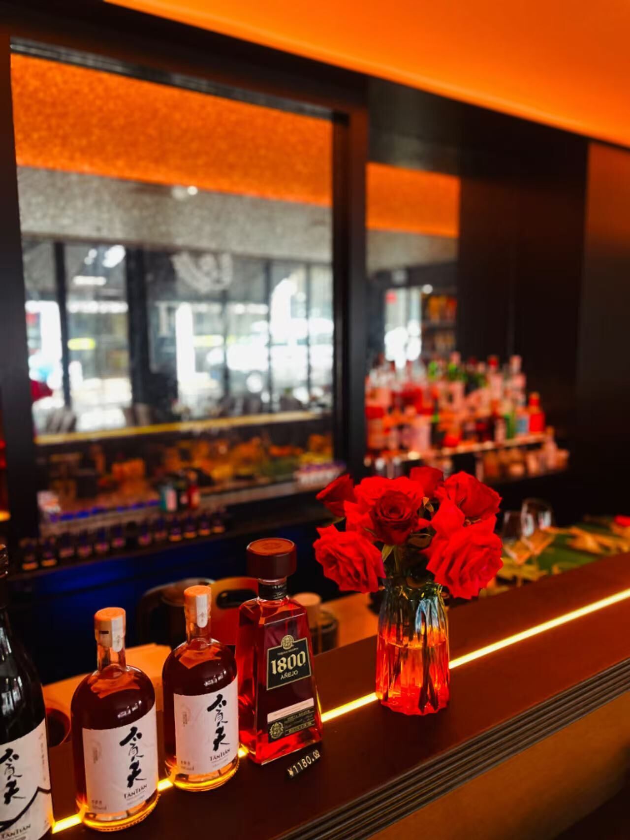 A bar area with bottles of liquor, a vase with red roses, and a reflection of the interior.