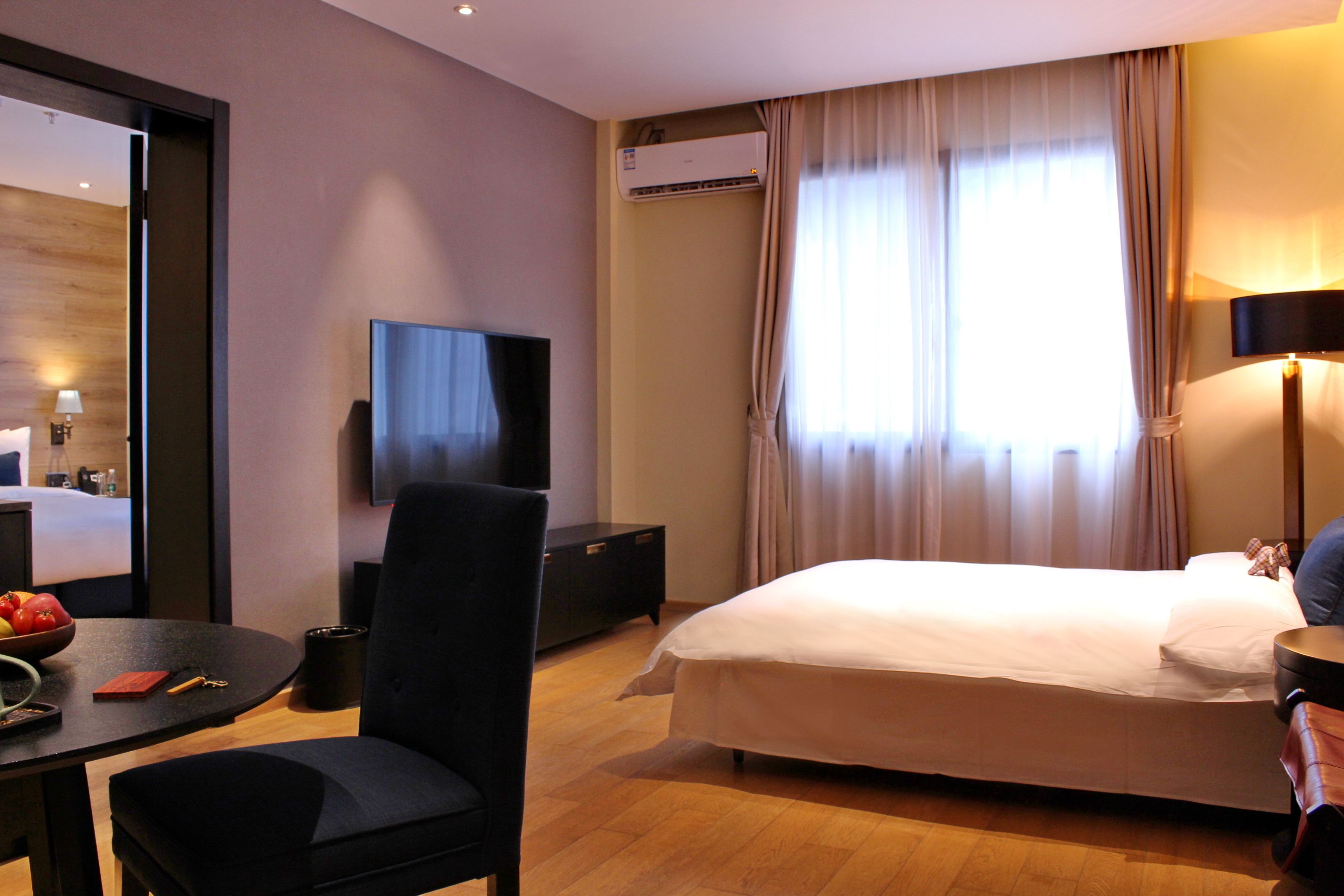 A hotel room with a bed, a TV, a desk with a chair, a bowl of fruit, and a window with curtains.