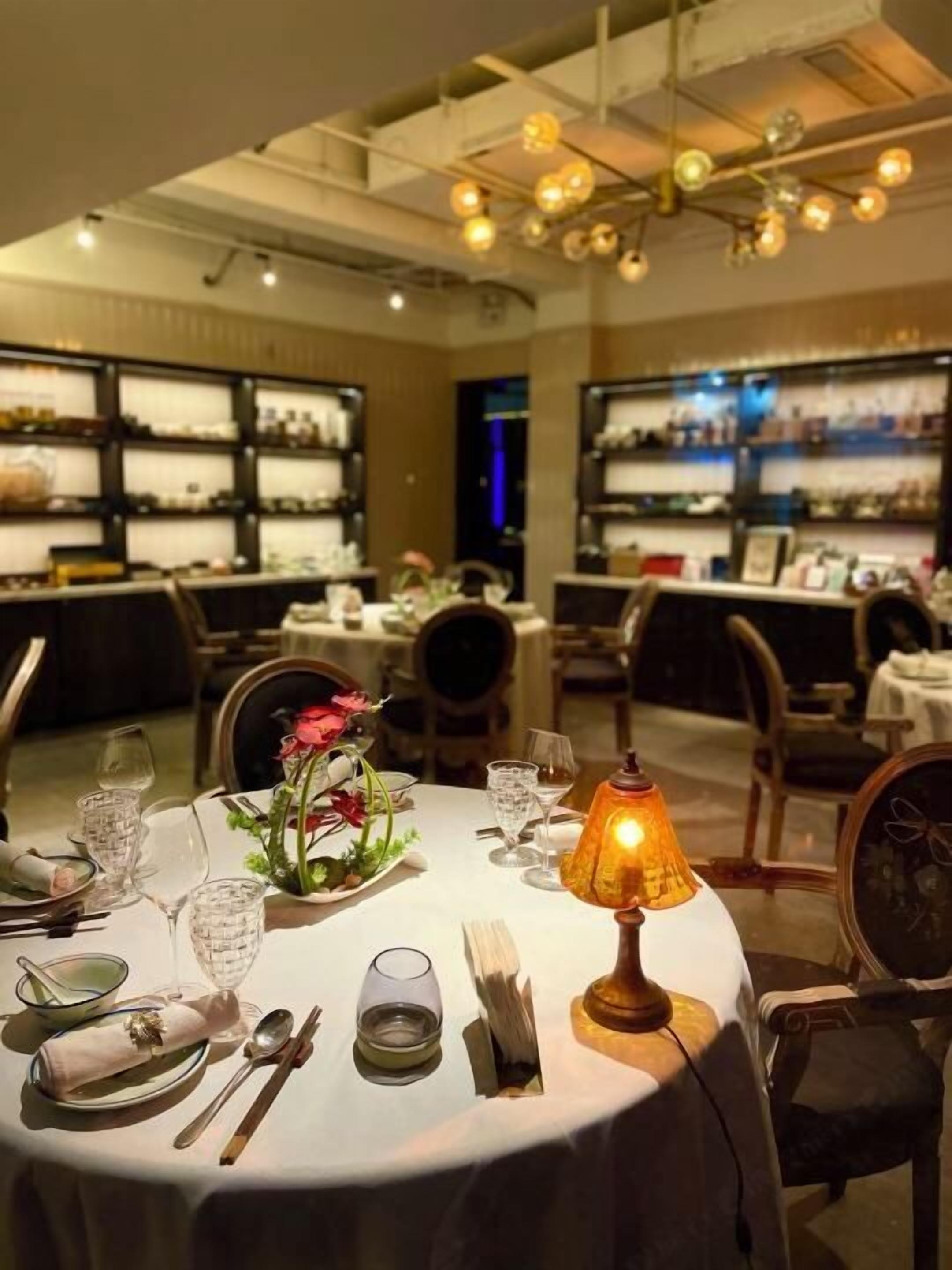 Een eetgedeelte met ronde tafels voor twee personen, met witte tafelkleden, glazen en bestek. Op tafel staat een warme lamp en een vaas met bloemen.