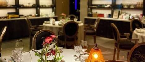 Un comedor con mesas redondas dispuestas para dos personas, con manteles blancos, cristalería y cubiertos. Una lámpara de mesa cálida y un jarrón con flores están sobre la mesa.